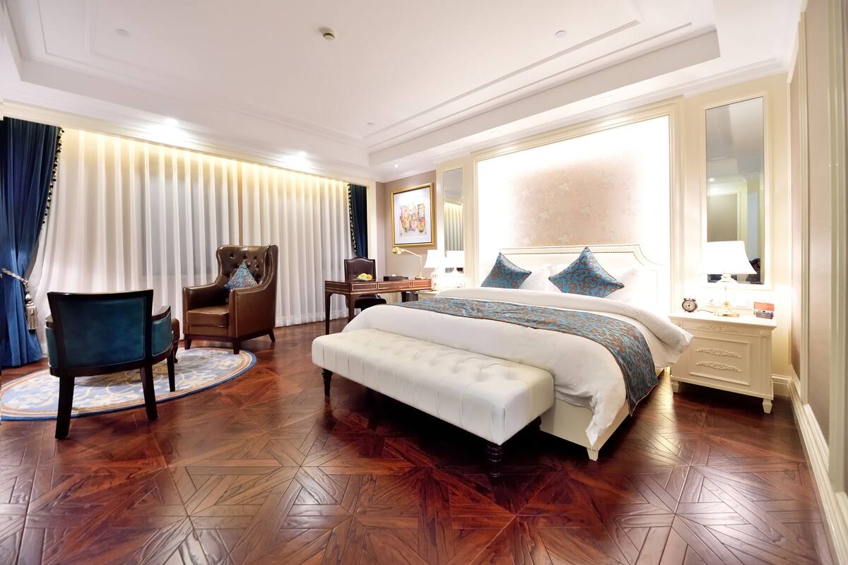 A spacious bedroom with a large bed, a sitting area with a chair and a bench, a desk, and a nightstand.