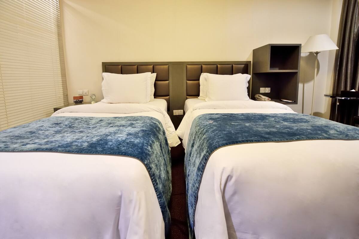 A hotel room with two single beds, a nightstand with a lamp, and a small shelf.