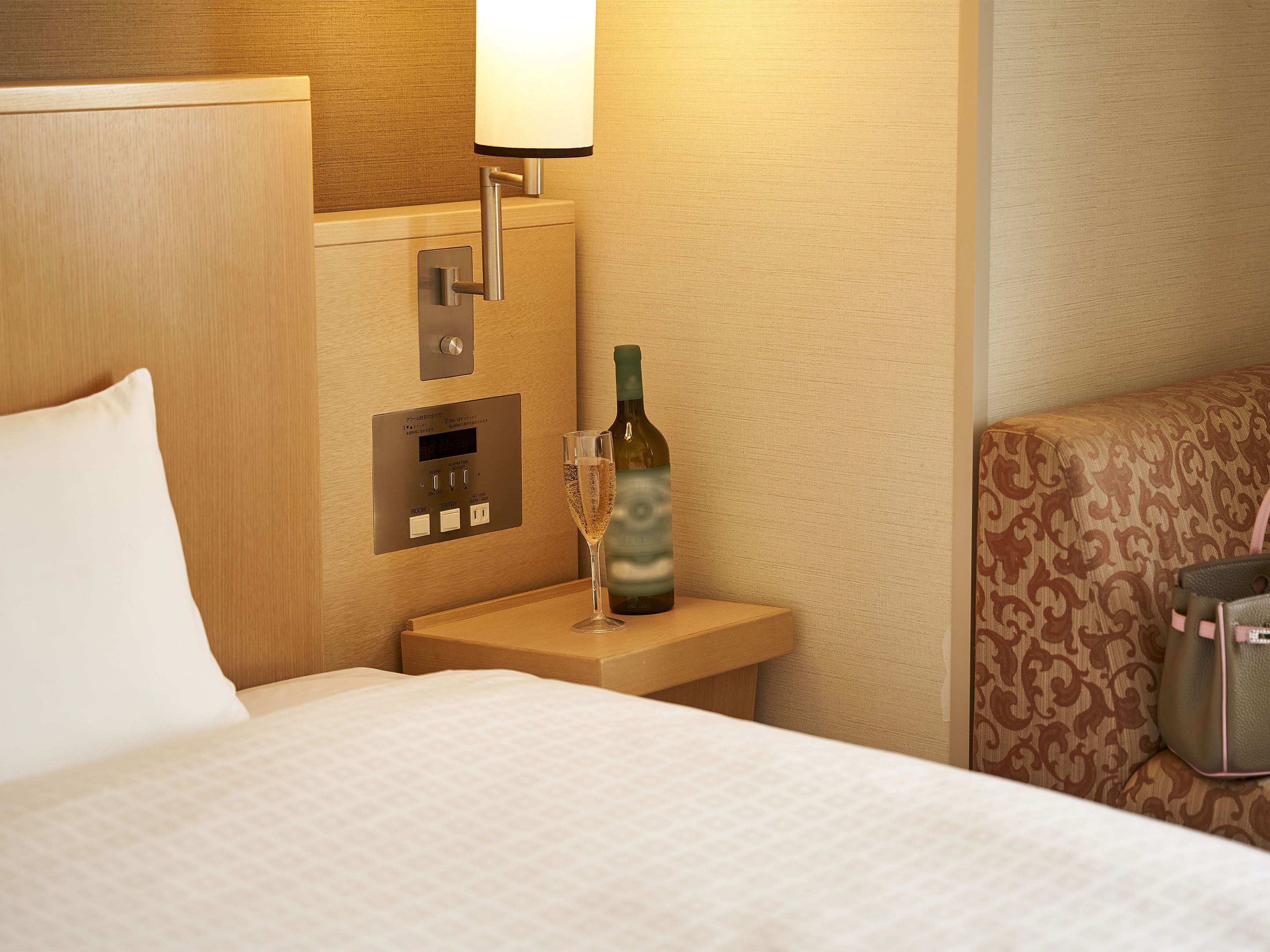 Une chambre d’hôtel avec un lit, une table de chevet sur laquelle se trouve une bouteille de vin, une lampe et un panneau de commande.