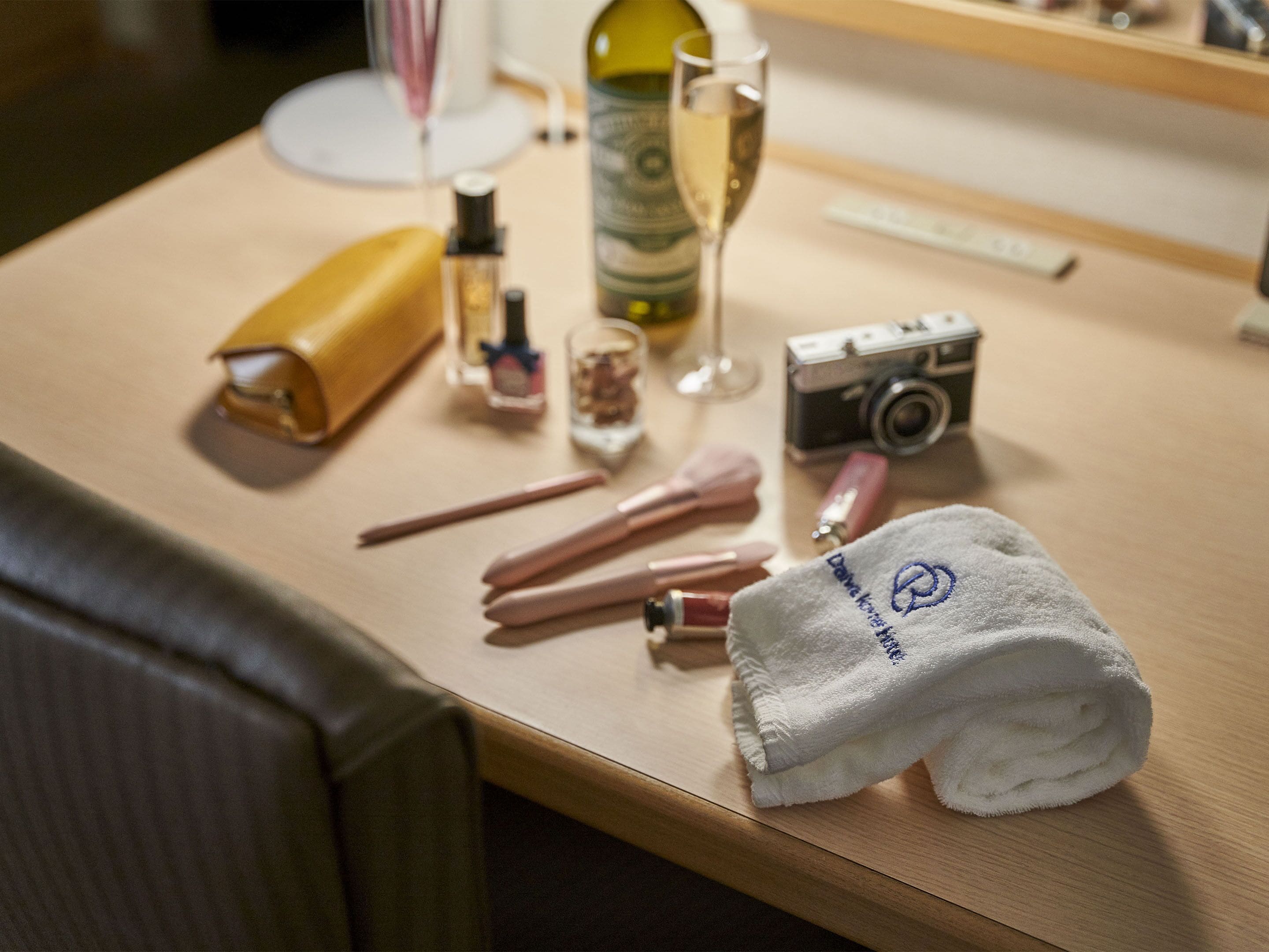 Une table bien rangée, avec une serviette, des pinceaux de maquillage, un appareil photo, une bouteille de vin et un flacon de parfum.