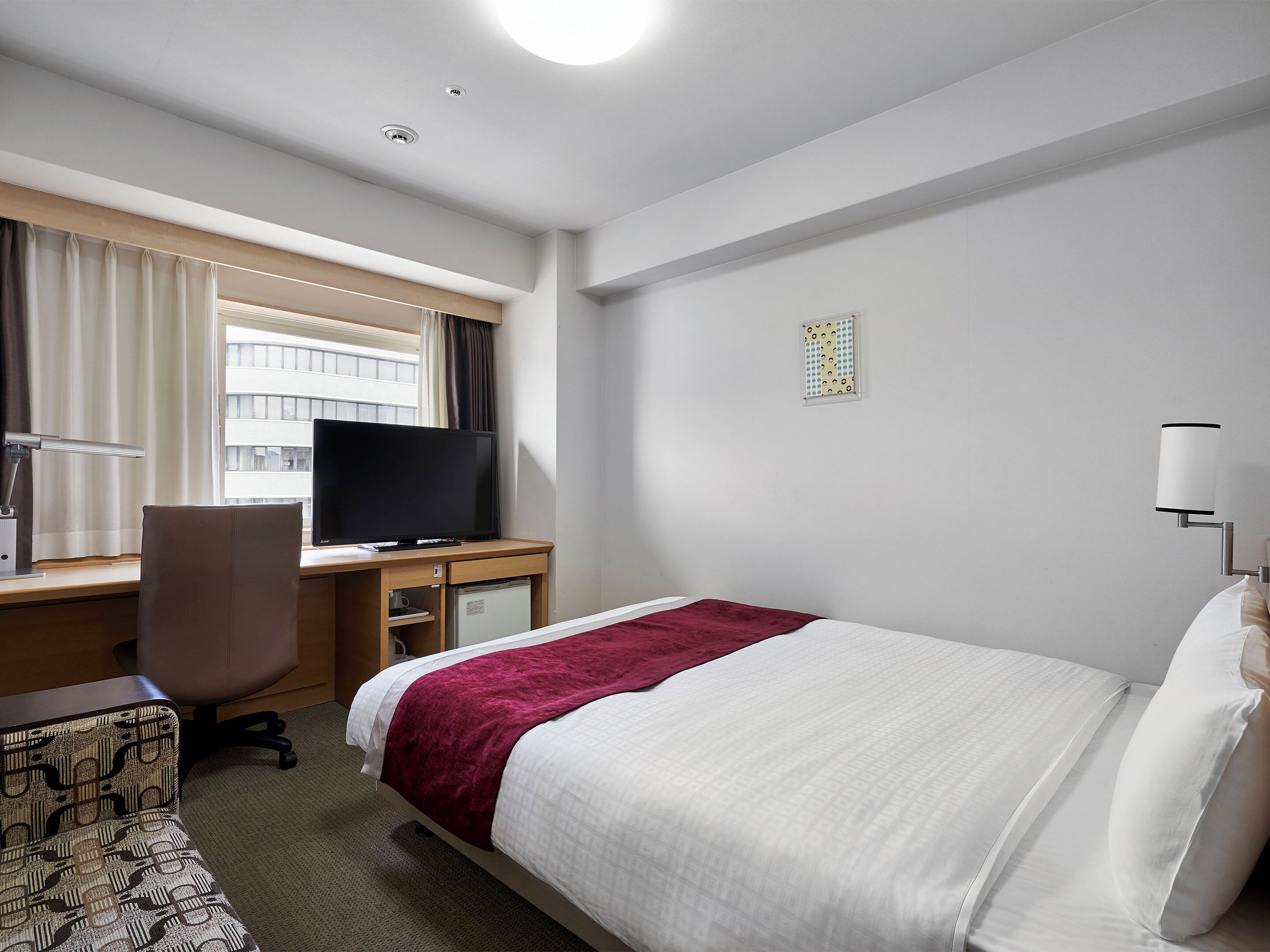 Une chambre d’hôtel avec un lit, un bureau équipé d’un ordinateur, une chaise, une télévision et une fenêtre avec des rideaux.