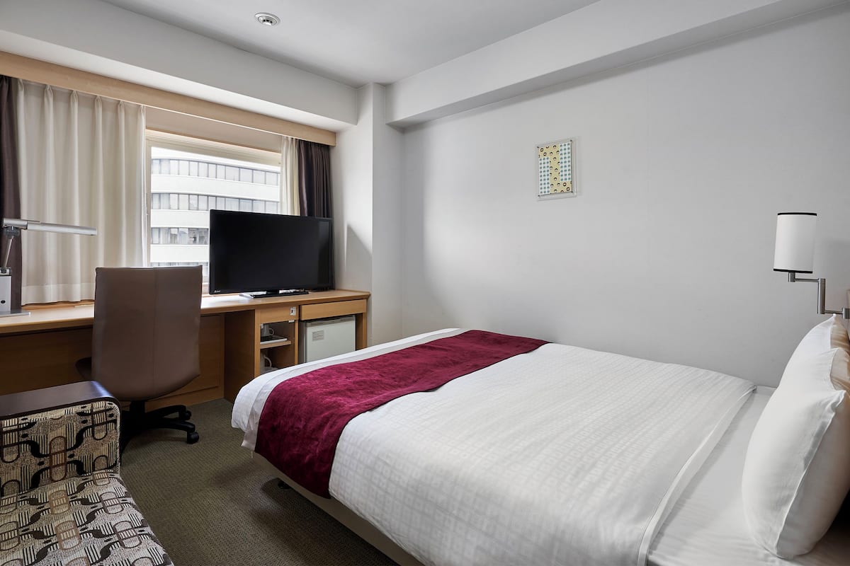 A hotel room with a bed, a desk with a computer, a chair, a television, and a window with curtains.