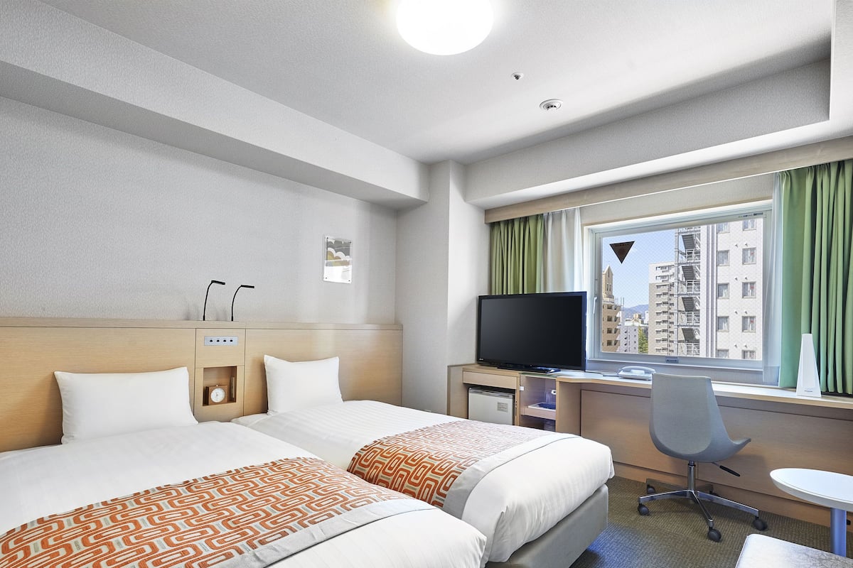 A hotel room with two beds, a desk with a computer, a TV, and a window with a view of buildings.