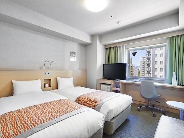 A hotel room with two beds, a desk with a computer, a TV, and a window with a view of buildings.
