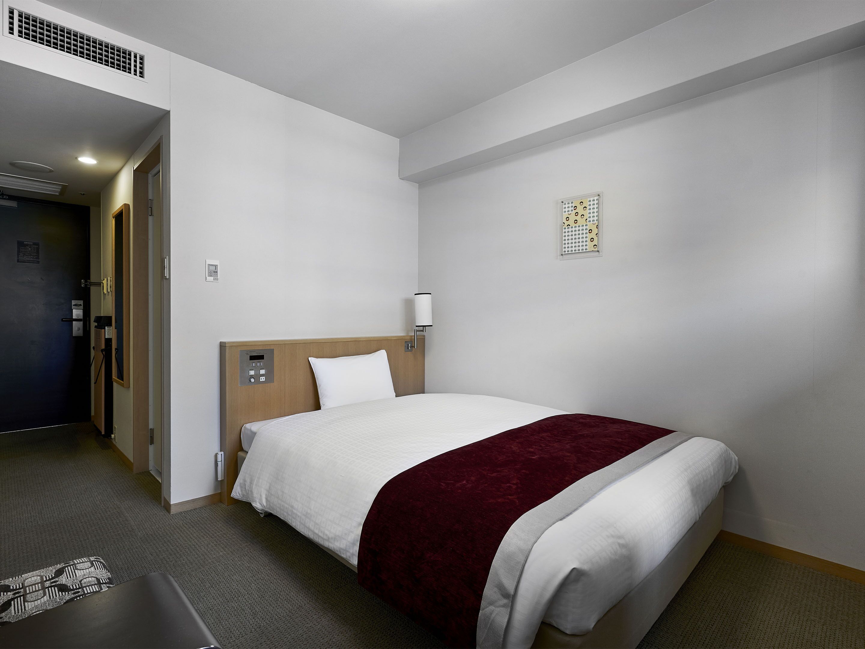 A hotel room with a bed, a headboard, a bedside table, a lamp, a wall-mounted air vent, and a door.