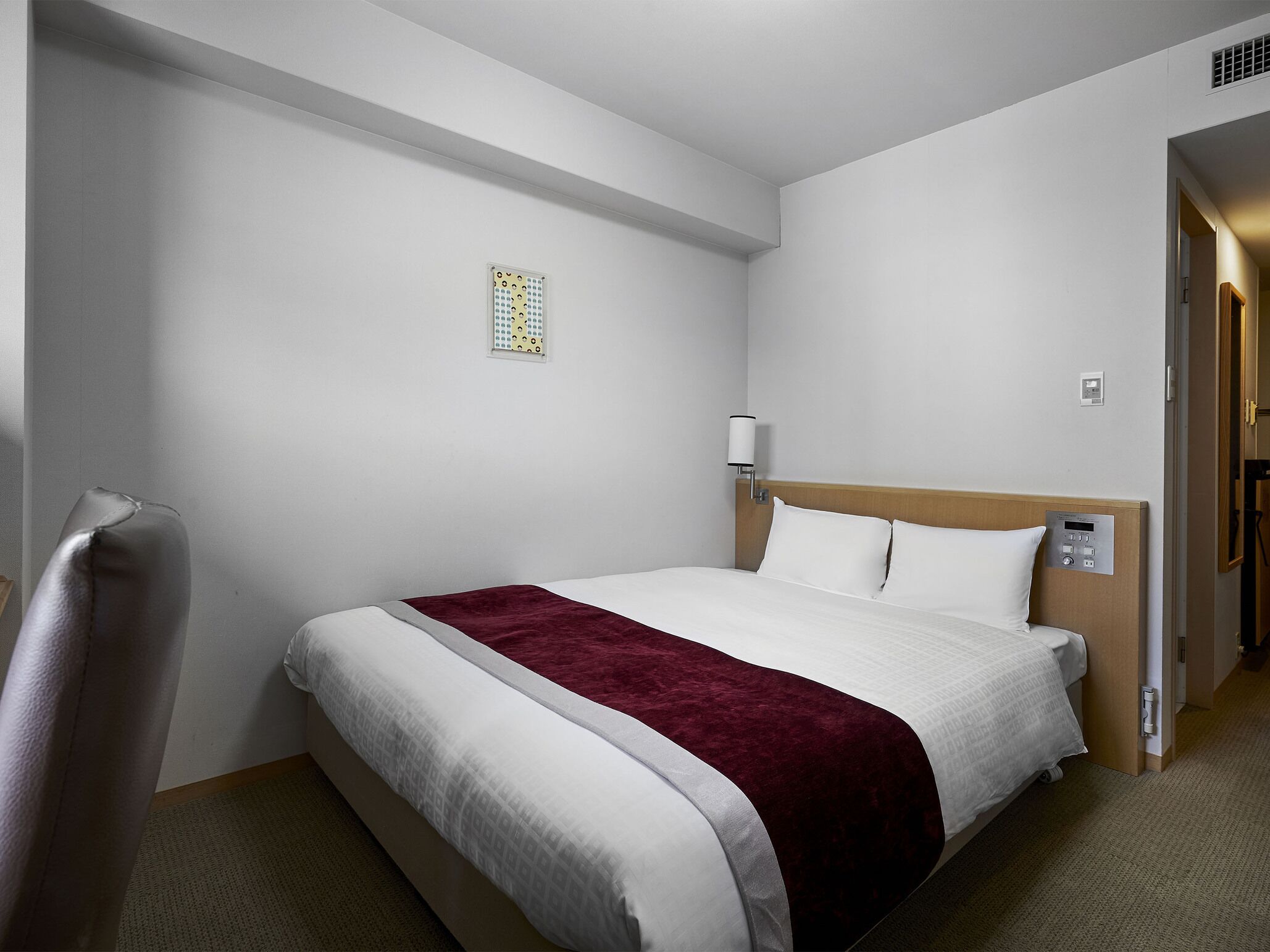 Een hotelkamer met een bed, nachtkastje, lamp, bedieningspaneel en een klein raam met decoratief patroon.