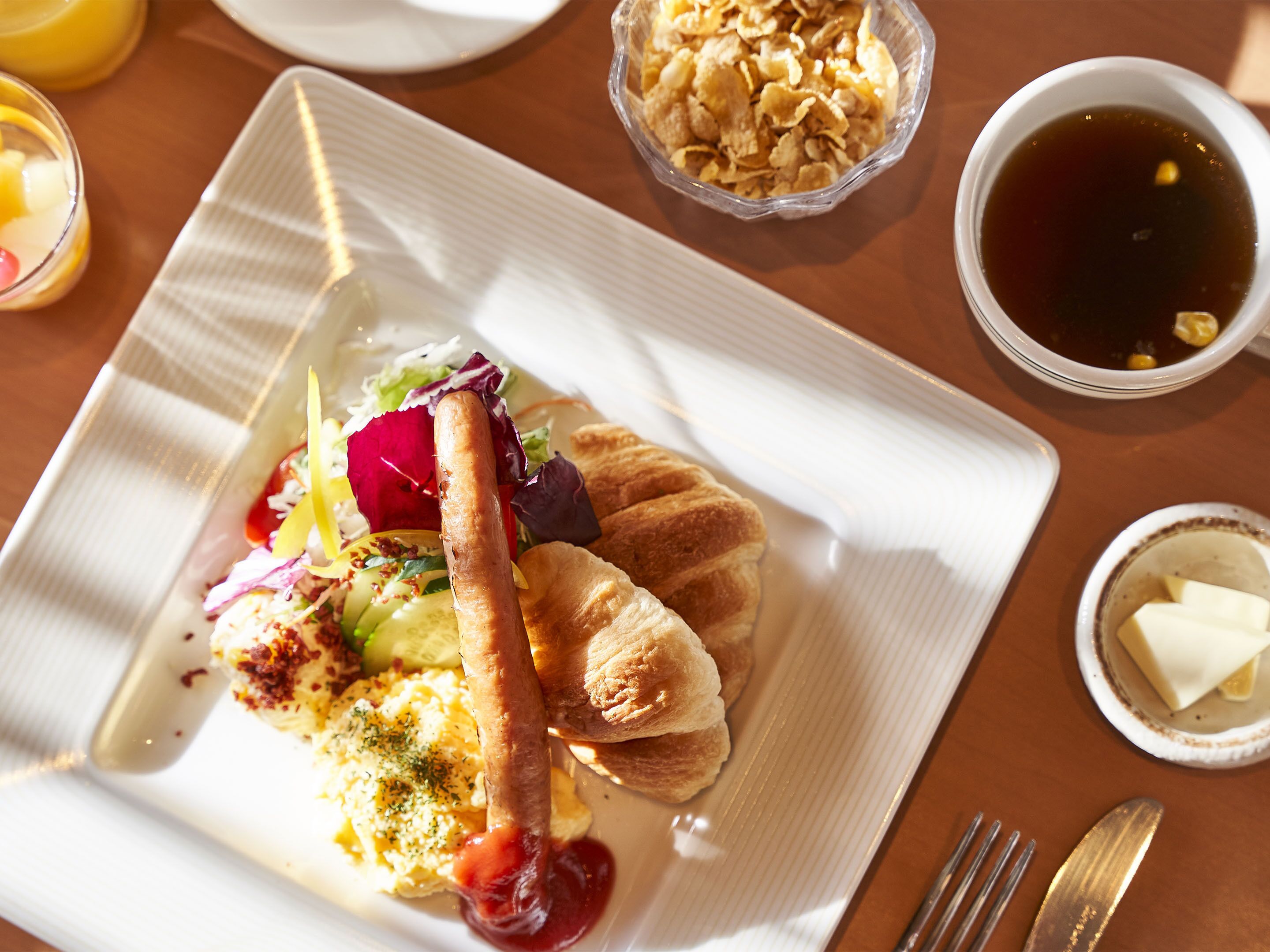 A meal with a croissant, a sausage, and a salad on a white plate.