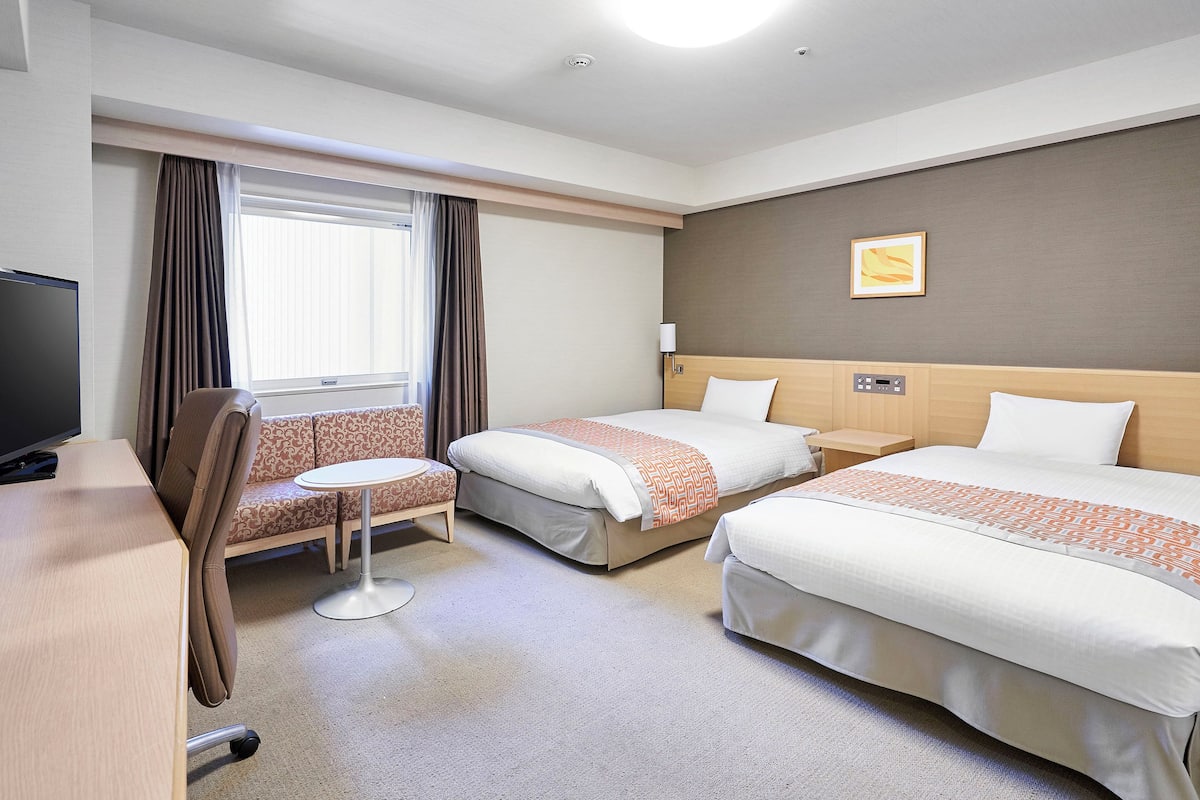 A hotel room with two beds, a desk, a chair, a small table, and a television.