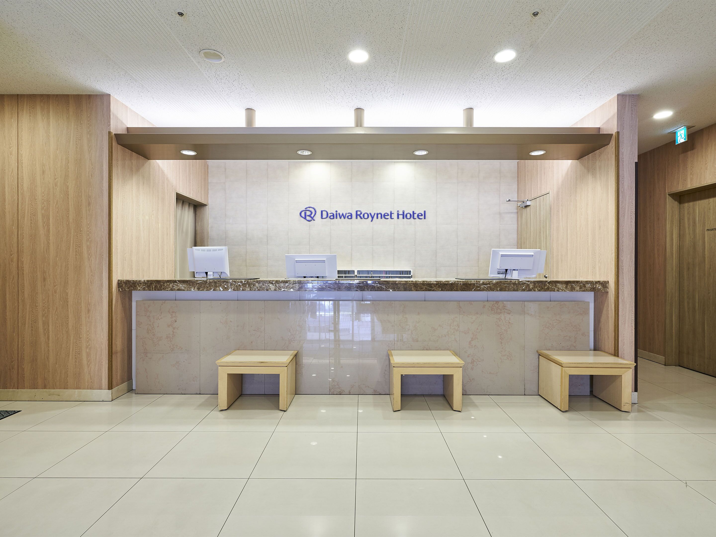 A reception desk at Daiwa Roynet Hotel with two computer monitors, a cash register, and two wooden stools.