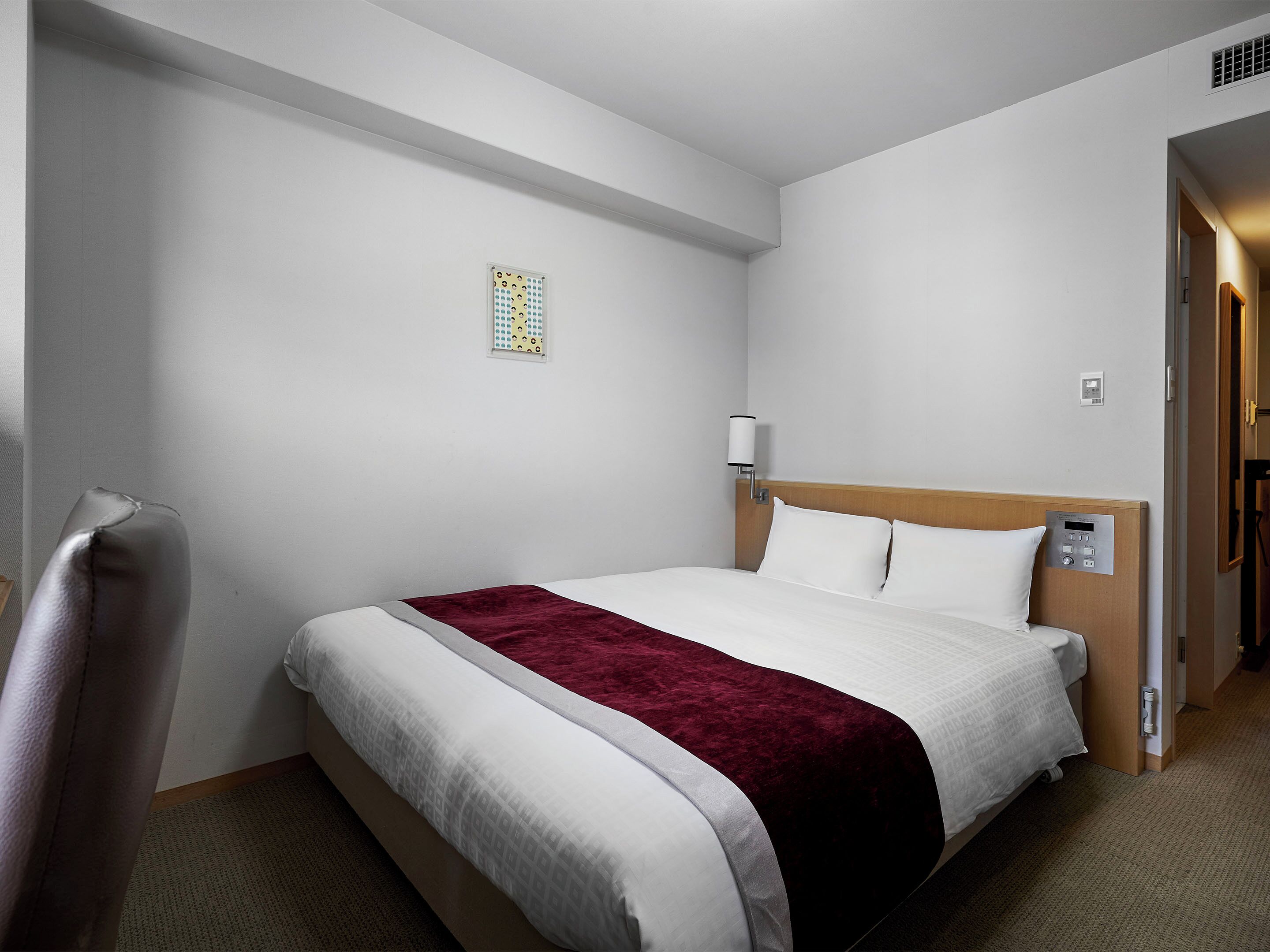 Une chambre d’hôtel avec un lit bien fait, une tête de lit dotée d’un panneau de commande, une table de chevet et une petite fenêtre ornée d’un motif décoratif.