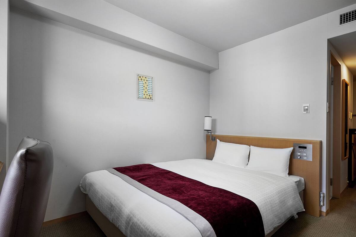 A hotel room with a neatly made bed, a headboard with a control panel, a bedside table, and a small window with a decorative pattern.