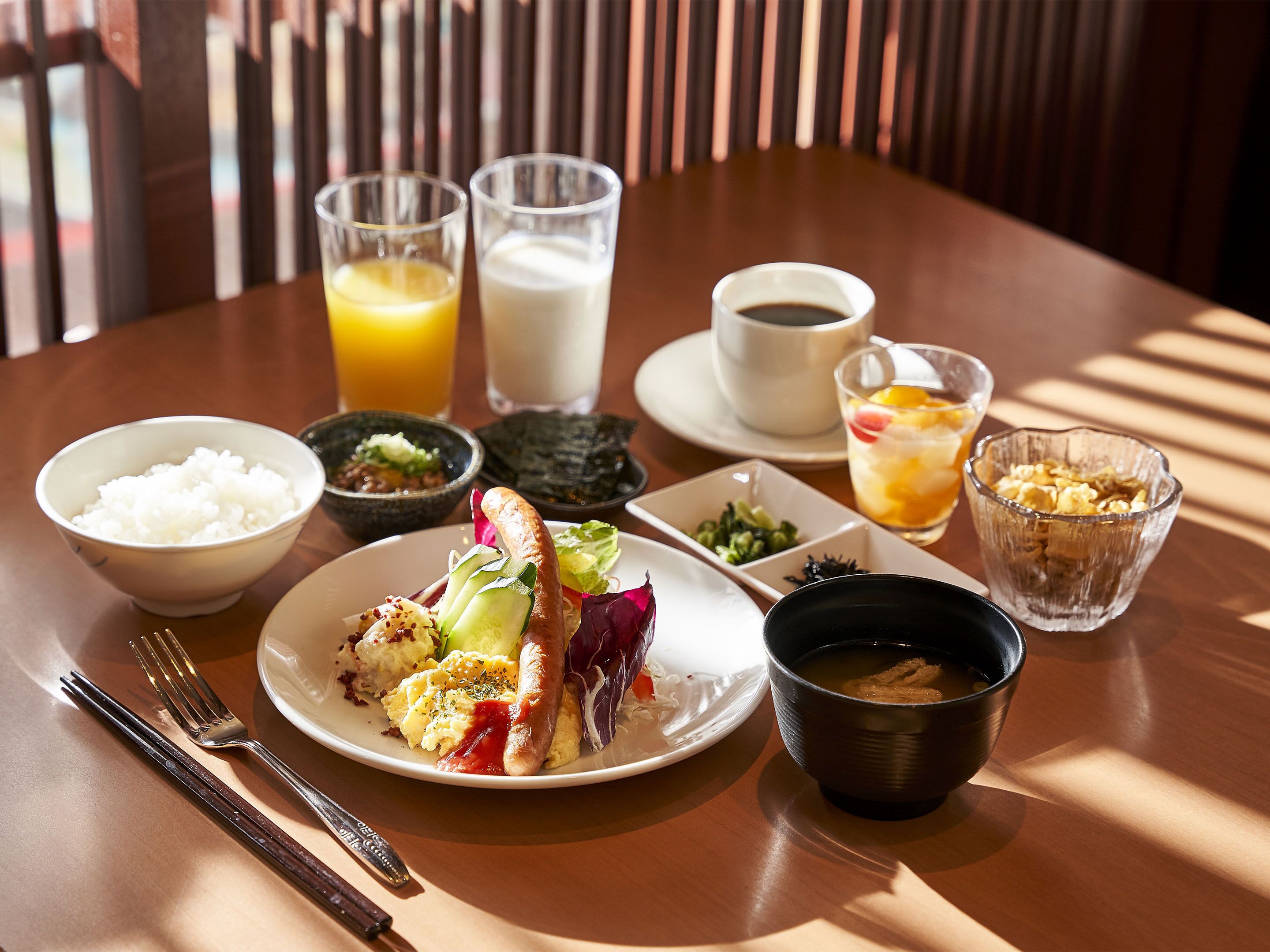 A dining table with a meal consisting of rice, vegetables, and a sausage, accompanied by drinks and a cup of coffee.