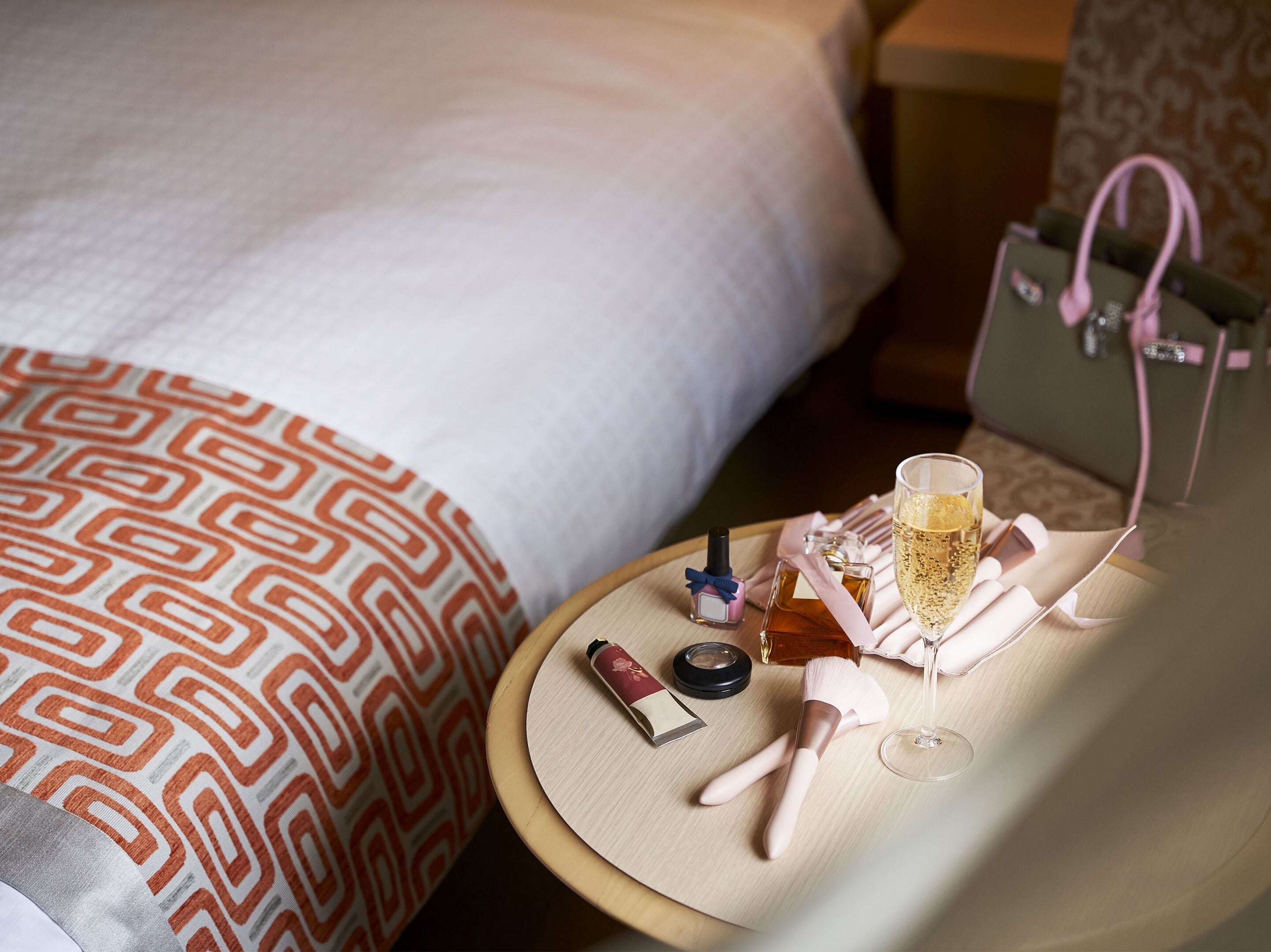 A neatly arranged bedside table with a bottle of perfume, a makeup brush, a compact, a glass of champagne, and a small handbag.