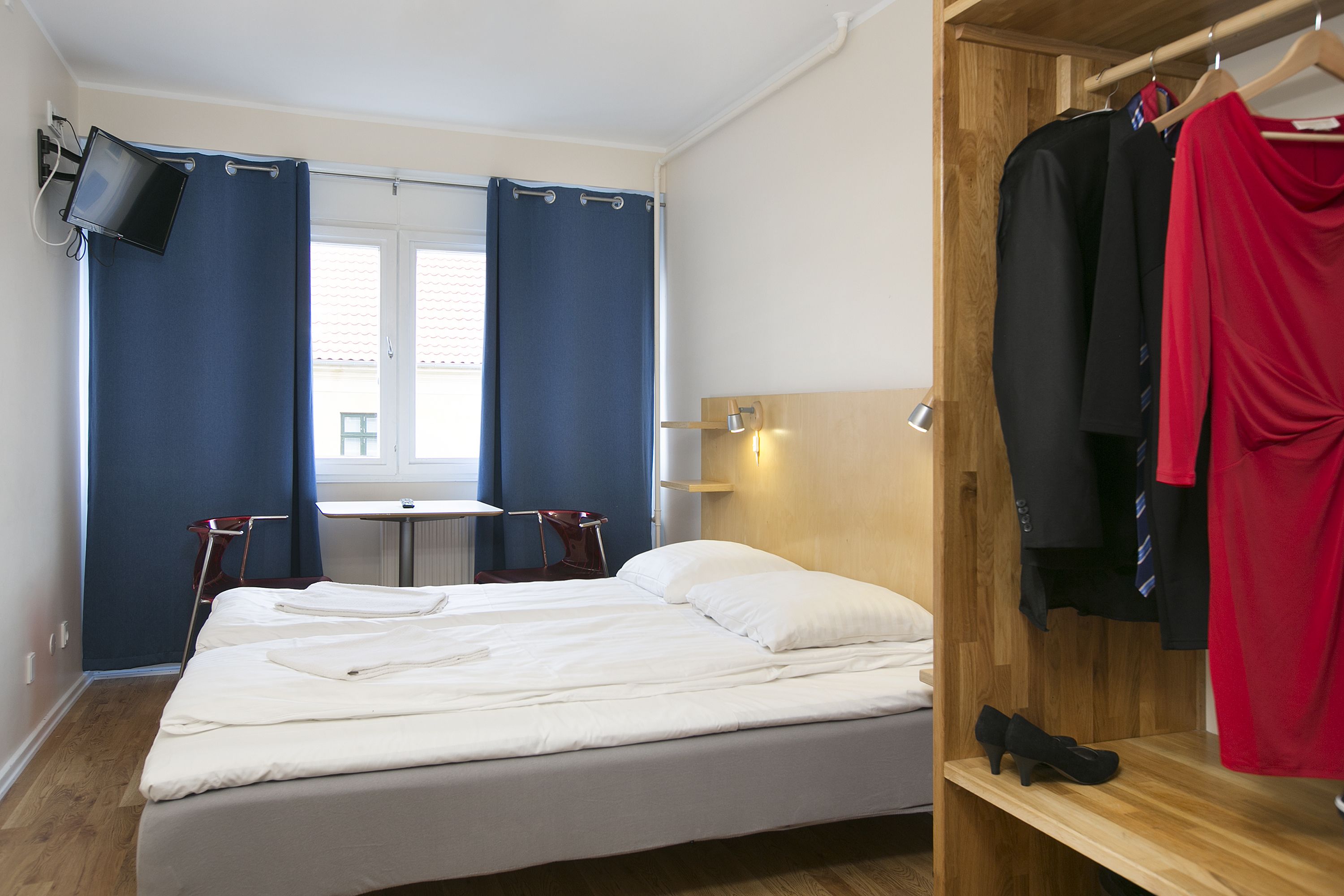 Quarto de hotel com uma cama, televisão fixada na parede, guarda-roupa de madeira e uma pequena mesa com cadeiras.