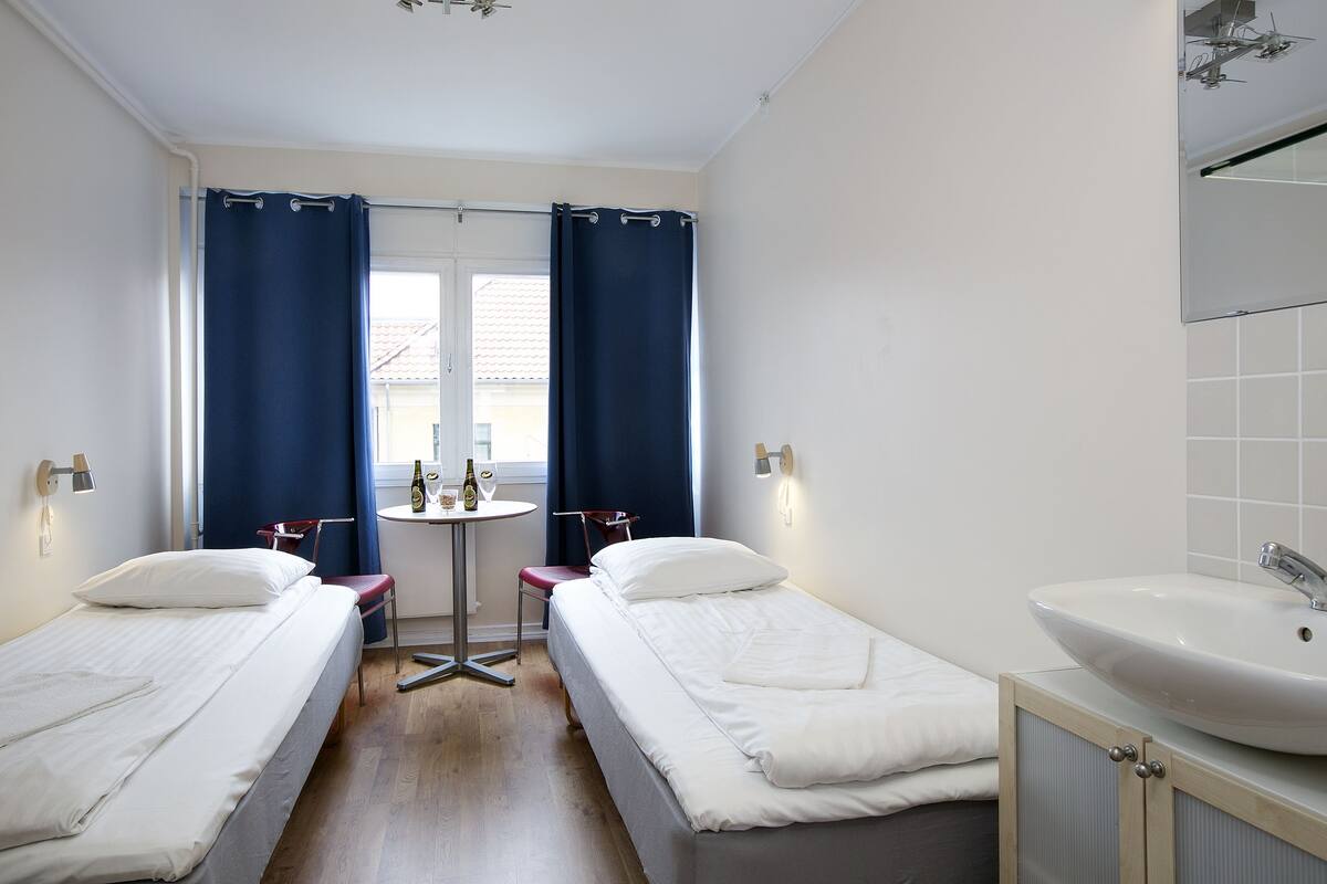 A hotel room with two single beds, a small table with chairs, a sink, and a window with curtains.