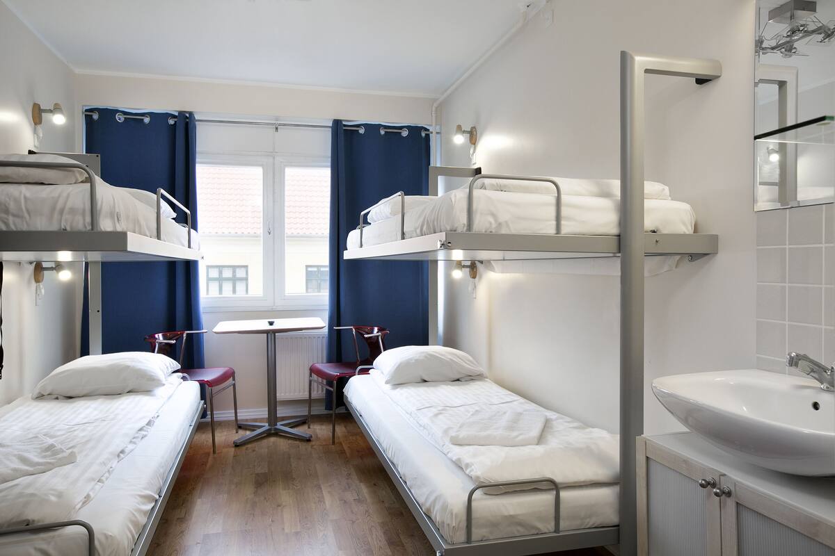 A dormitory room with bunk beds, a small table, a chair, a sink, and a window with curtains.