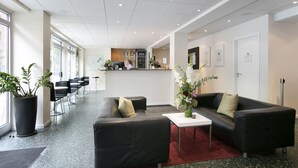 Un moderno vestíbulo de hotel con mostrador de recepción, zona de estar con sofás y sillones, plantas en macetas y un ventanal que proporciona luz natural.