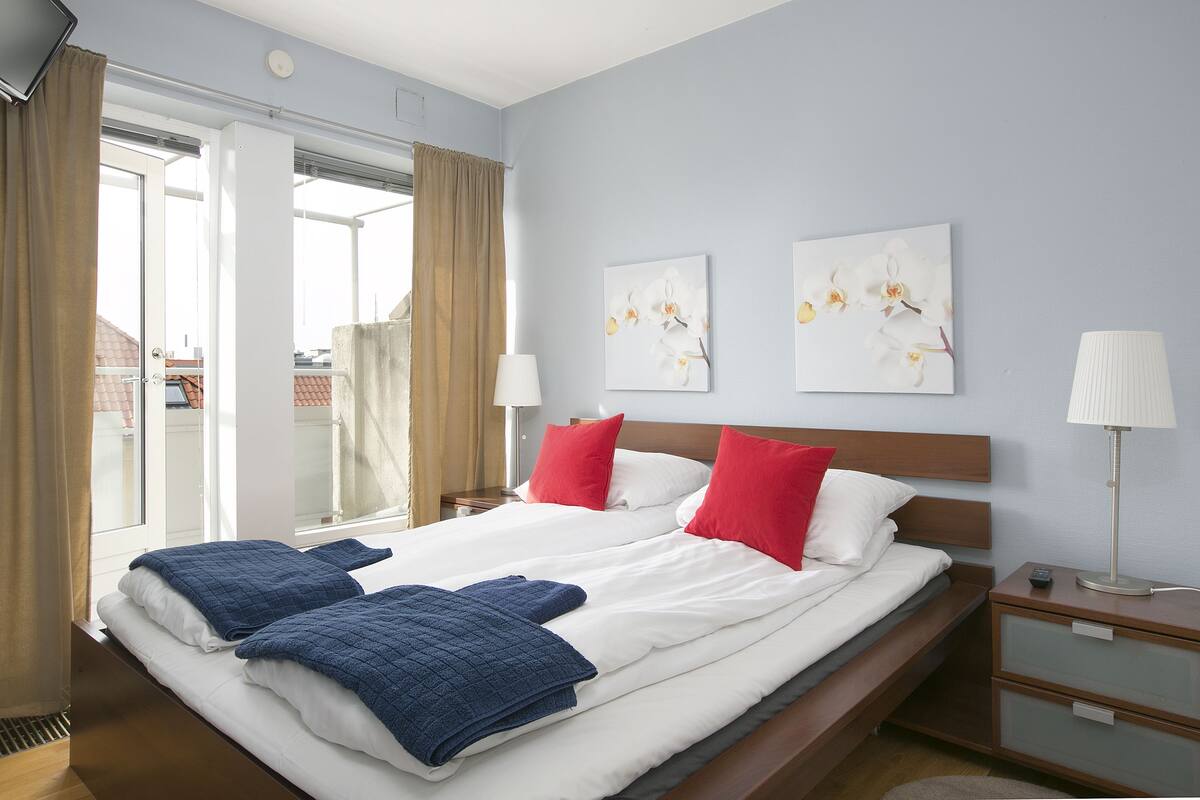 A double bed with white and blue bedding, red pillows, and a nightstand with a lamp.