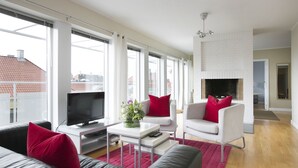 Una sala de estar moderna con un televisor de pantalla plana, una mesa de centro blanca, un sofá y dos sillones con cojines rojos. Hay una chimenea con detalles de ladrillo blanco y un ventanal con vistas a edificios.