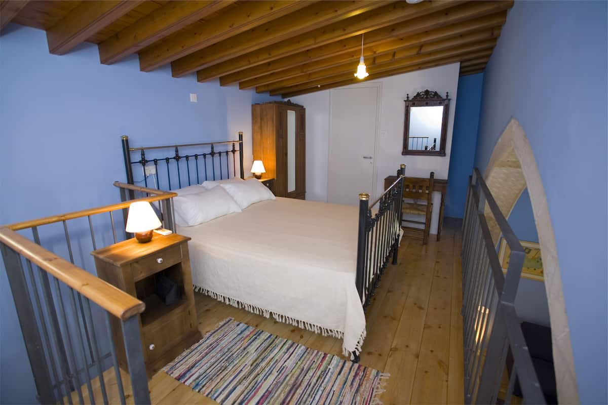 A bedroom with a wooden ceiling, a bed with a white bedspread, a nightstand with a lamp, a wooden wardrobe, and a mirror on the wall.