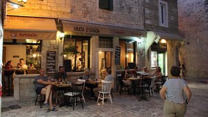Un café al aire libre con mesas y sillas, un letrero que dice 'VRATA O'GRADA' y personas cenando.
