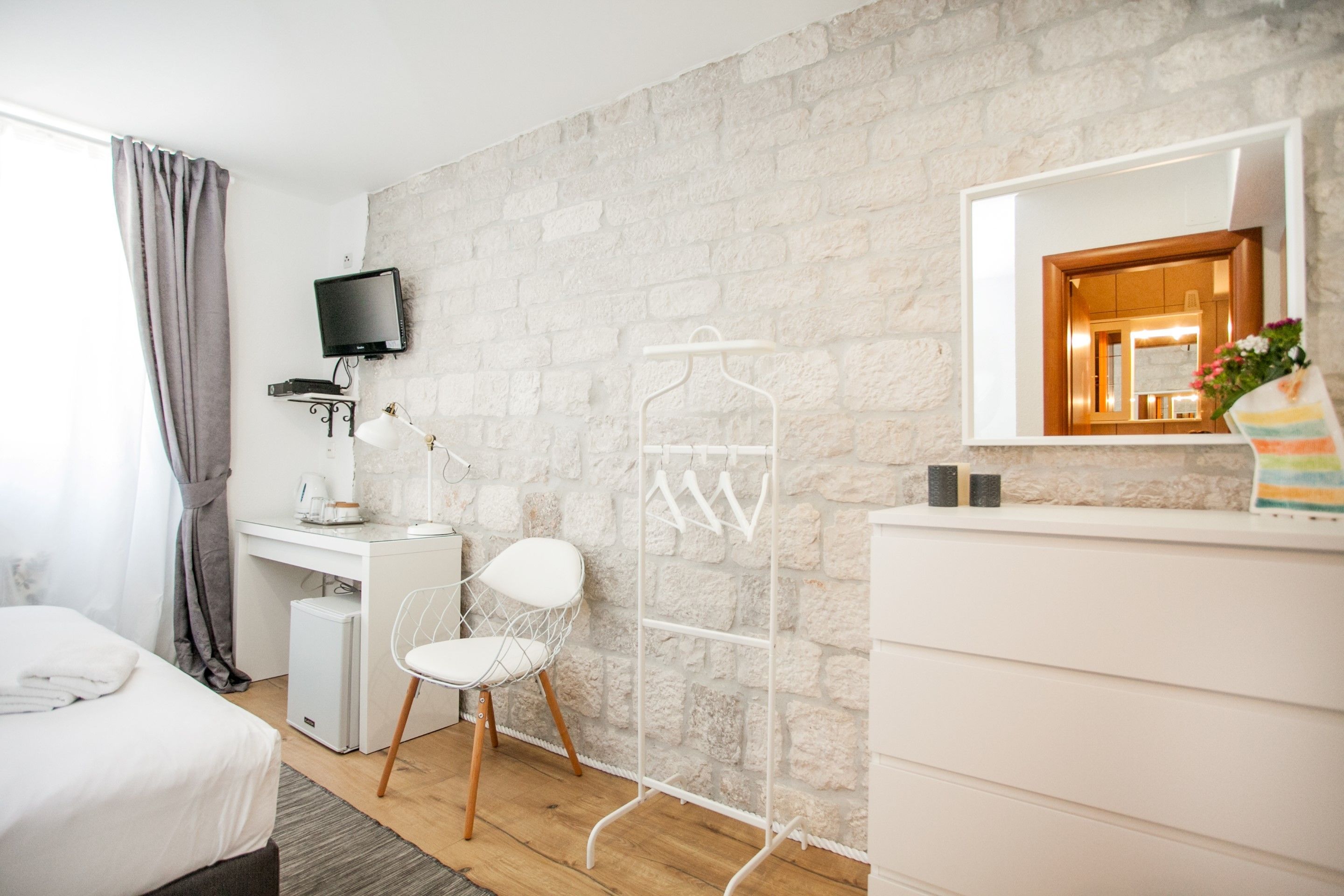 A modern hotel room with a stone wall, a flat-screen TV, a desk with a chair, a small fridge, a bed, and a wardrobe.