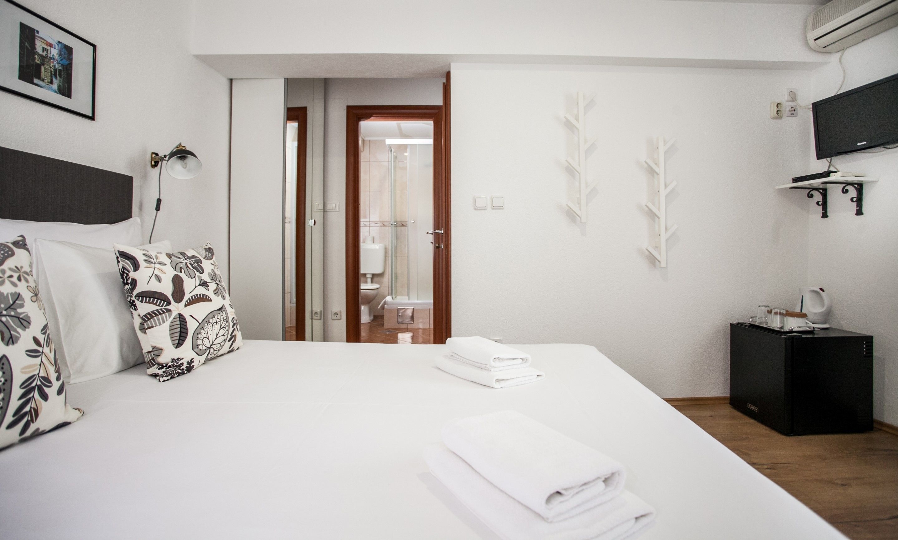 Quarto de hotel moderno com uma cama grande, mesa de cabeceira, televisão e área de chuveiro.