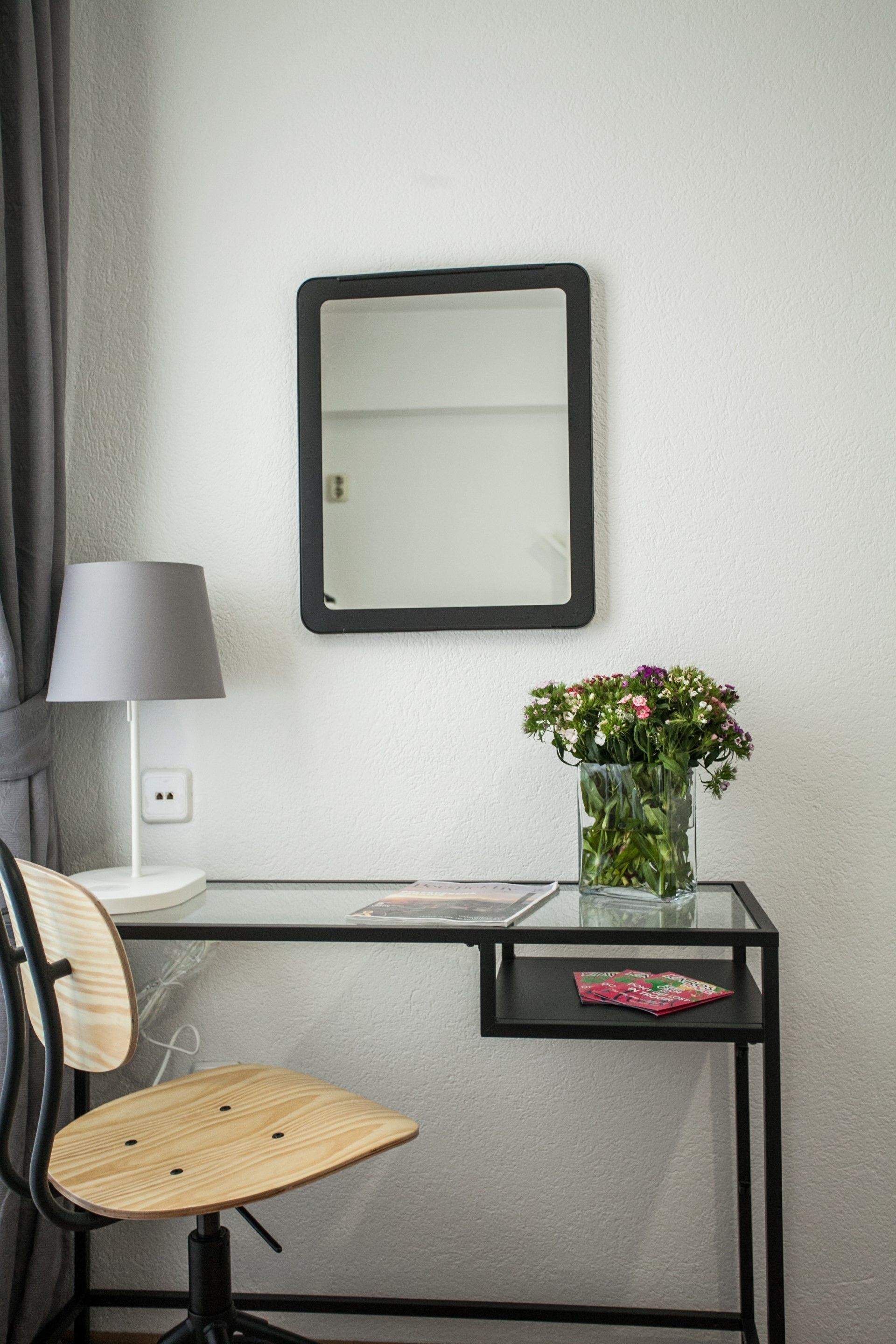 A modern desk with a chair, a lamp, a vase with flowers, and a magazine on a glass table.