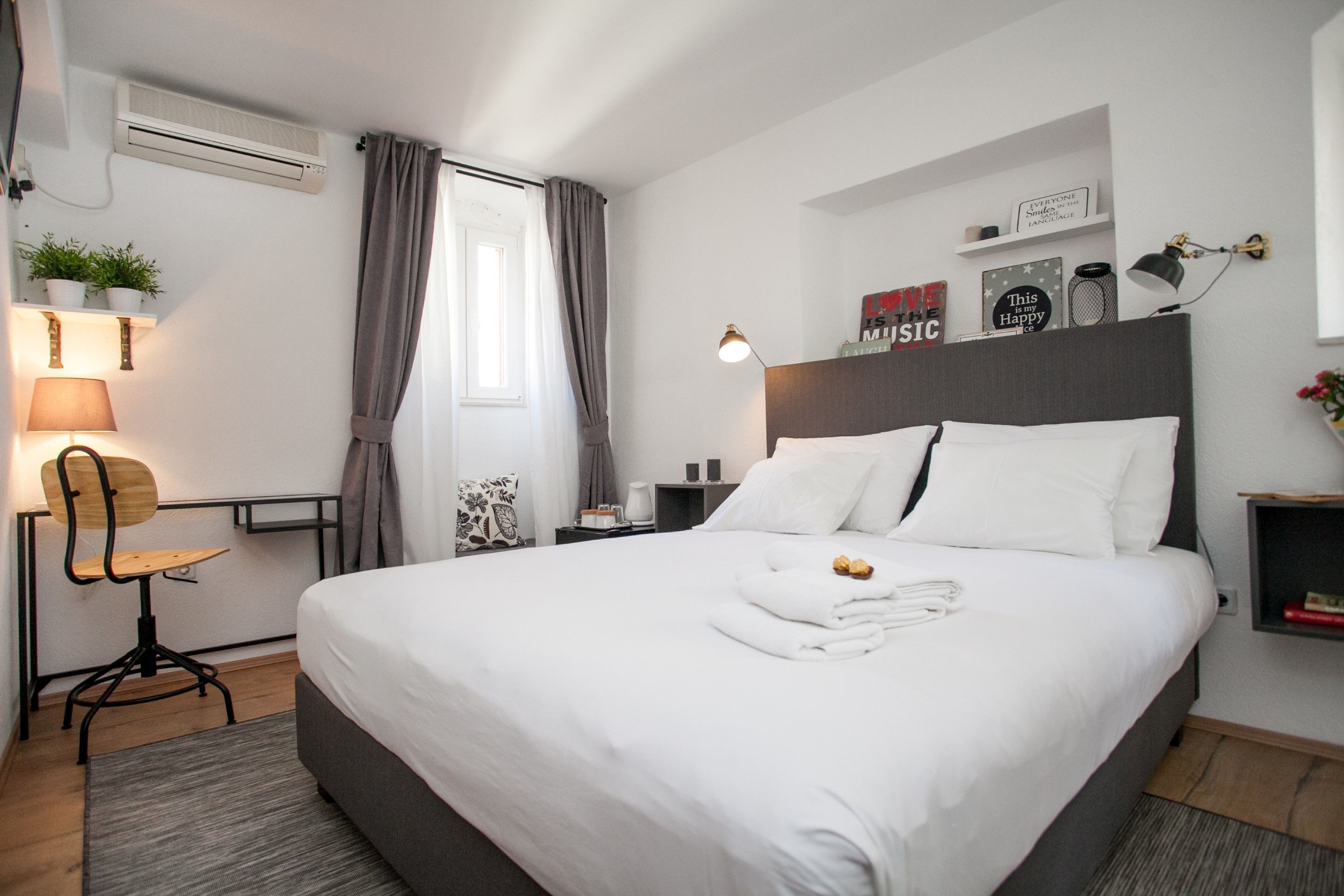 A modern hotel room with a large bed, a desk with a chair, a lamp, and a small shelf with decorative items.