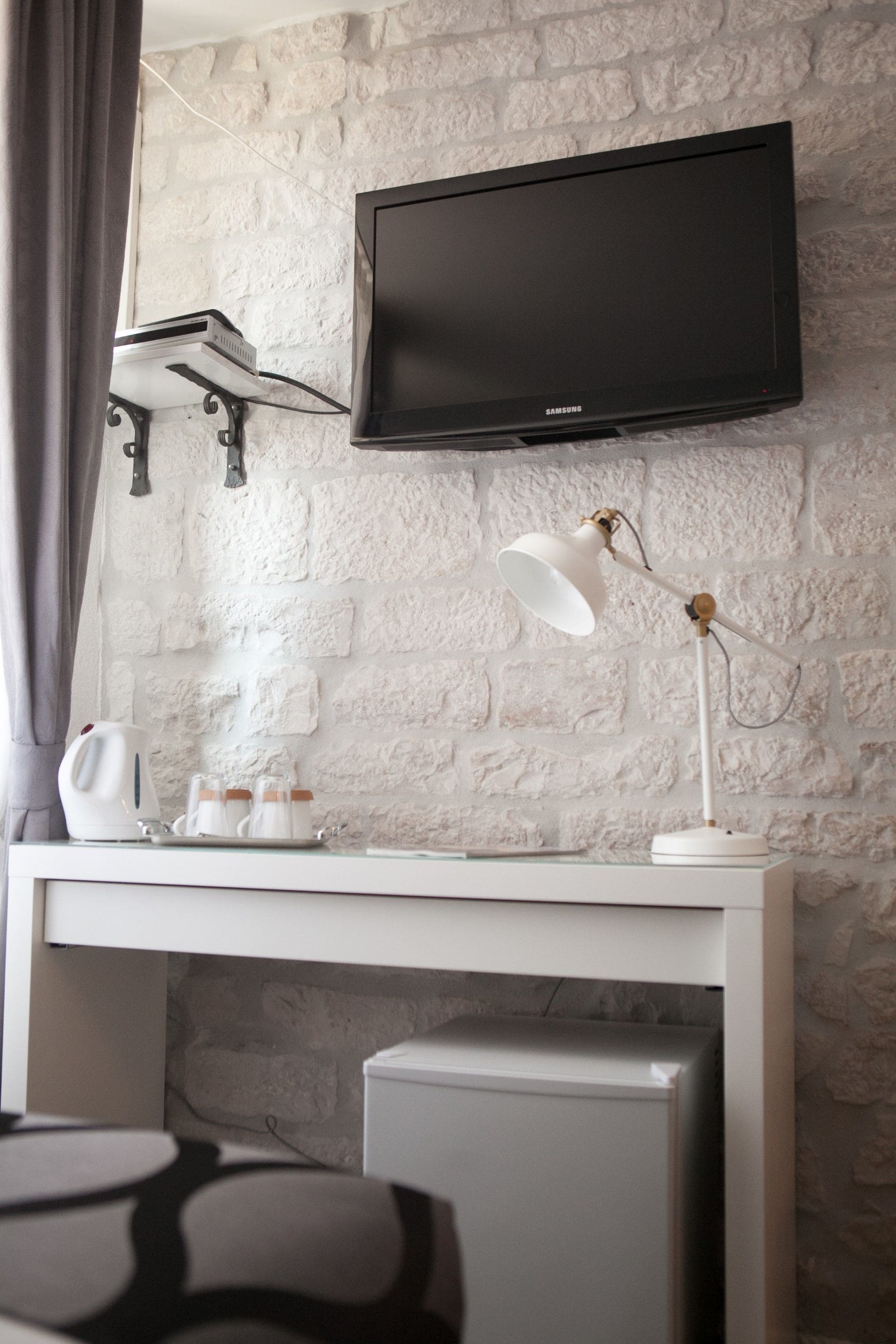 Et moderne værelse med en hvid murstensvæg, et vægmonteret fladskærms-tv, et hvidt skrivebord med lampe, elkedel og et lille køleskab.