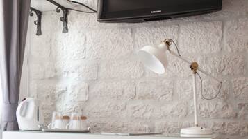 Une chambre moderne avec un mur en briques blanches, un téléviseur à écran plat fixé au mur, un bureau blanc avec une lampe, une bouilloire et un petit réfrigérateur.