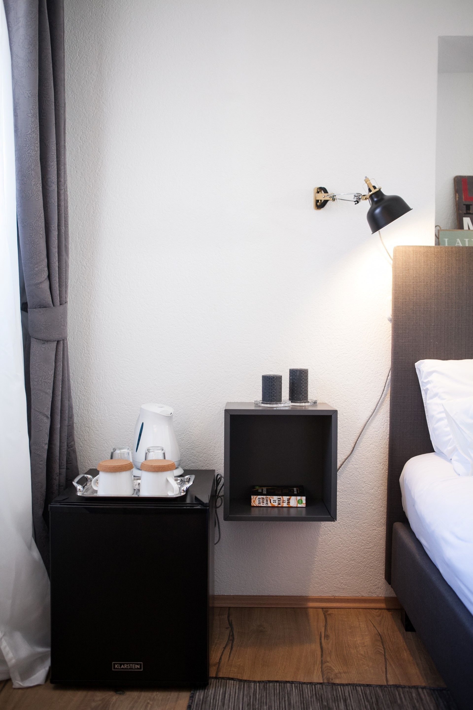 Une chambre d’hôtel avec un lit, une table de chevet avec une lampe, une petite étagère avec des livres et une machine à café.