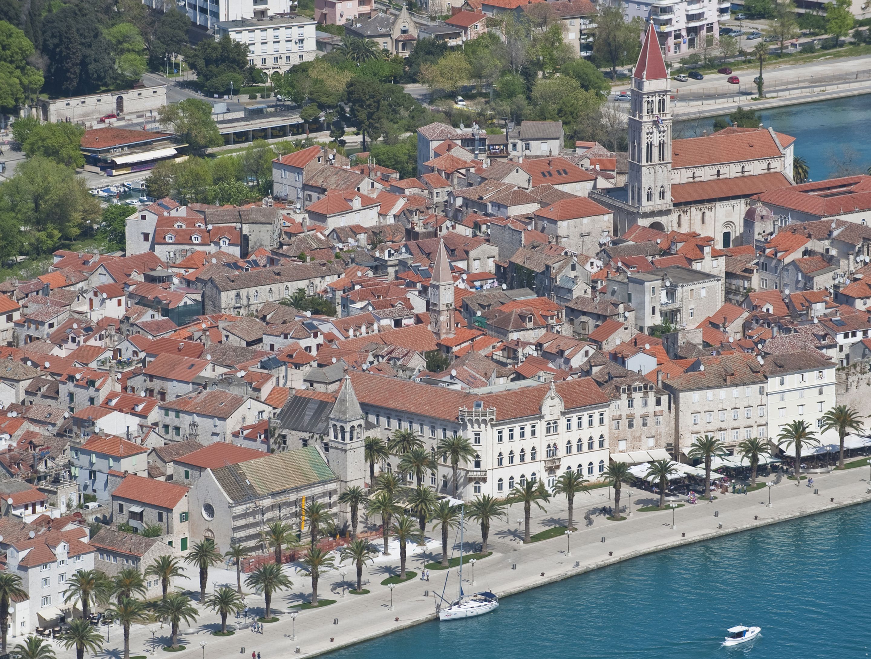 Vista aérea de un pueblo histórico con edificios apretados, una iglesia destacada con una torre alta y un paseo marítimo con palmeras.