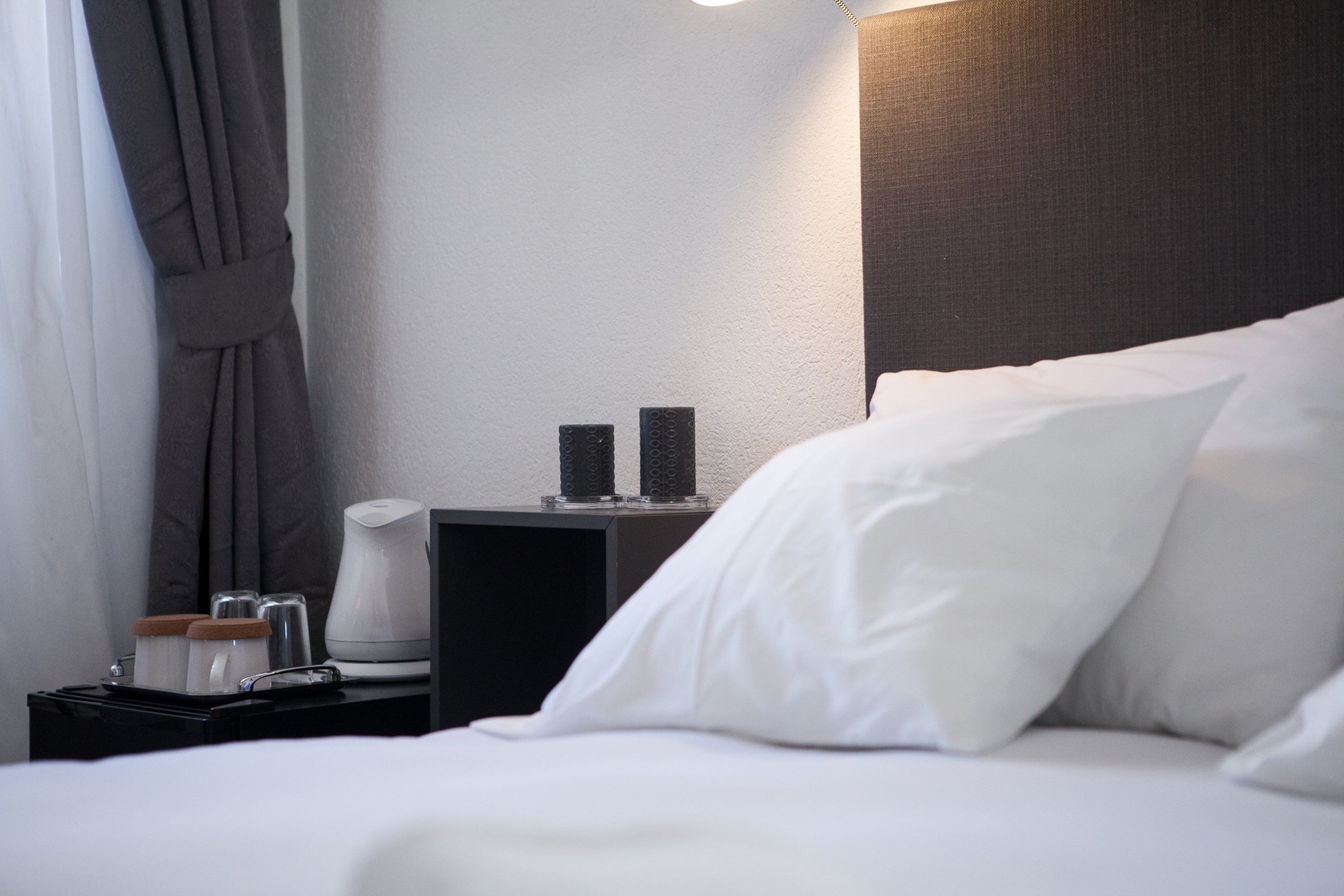 A neatly made bed with white linens, a bedside table with a lamp, and a small nightstand with a coffee maker and cups.
