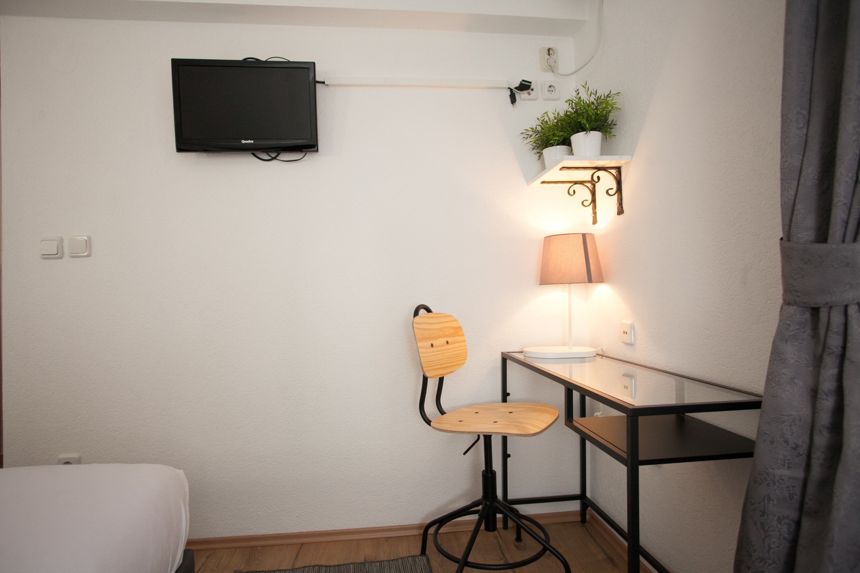 Una habitación pequeña y moderna con una cama individual, un escritorio con silla, un televisor montado en la pared y una lámpara de pared.