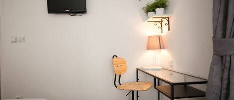 Una habitación pequeña y moderna con una cama individual, un escritorio con silla, un televisor montado en la pared y una lámpara de pared.