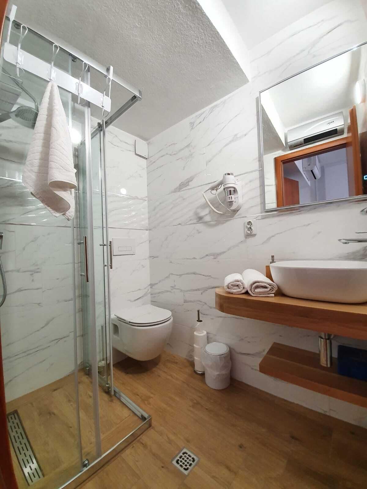 A modern bathroom with a glass shower enclosure, a white toilet, a wooden vanity with a sink, and a marble backsplash.