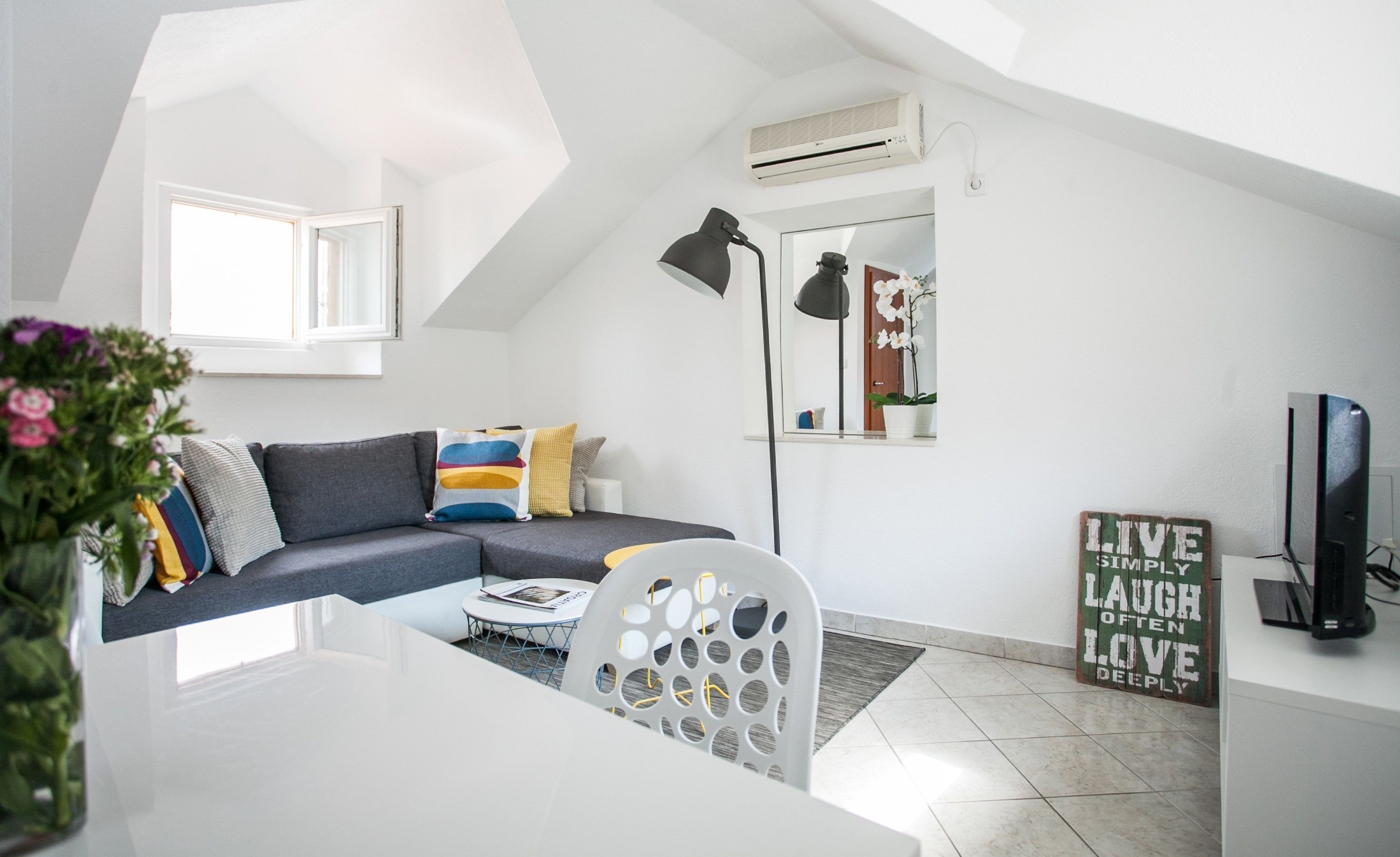 A modern living room with a grey sofa, a white coffee table, a black floor lamp, and a TV. There's a potted orchid on the windowsill and a sign with a motivational message.