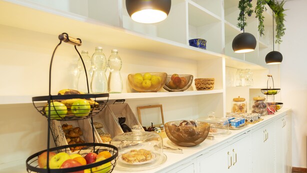 A buffet setup with various food items and containers, including baskets of fruits and bowls of food, on a white counter with shelves above. - Hotel Madrisol (Madrid)