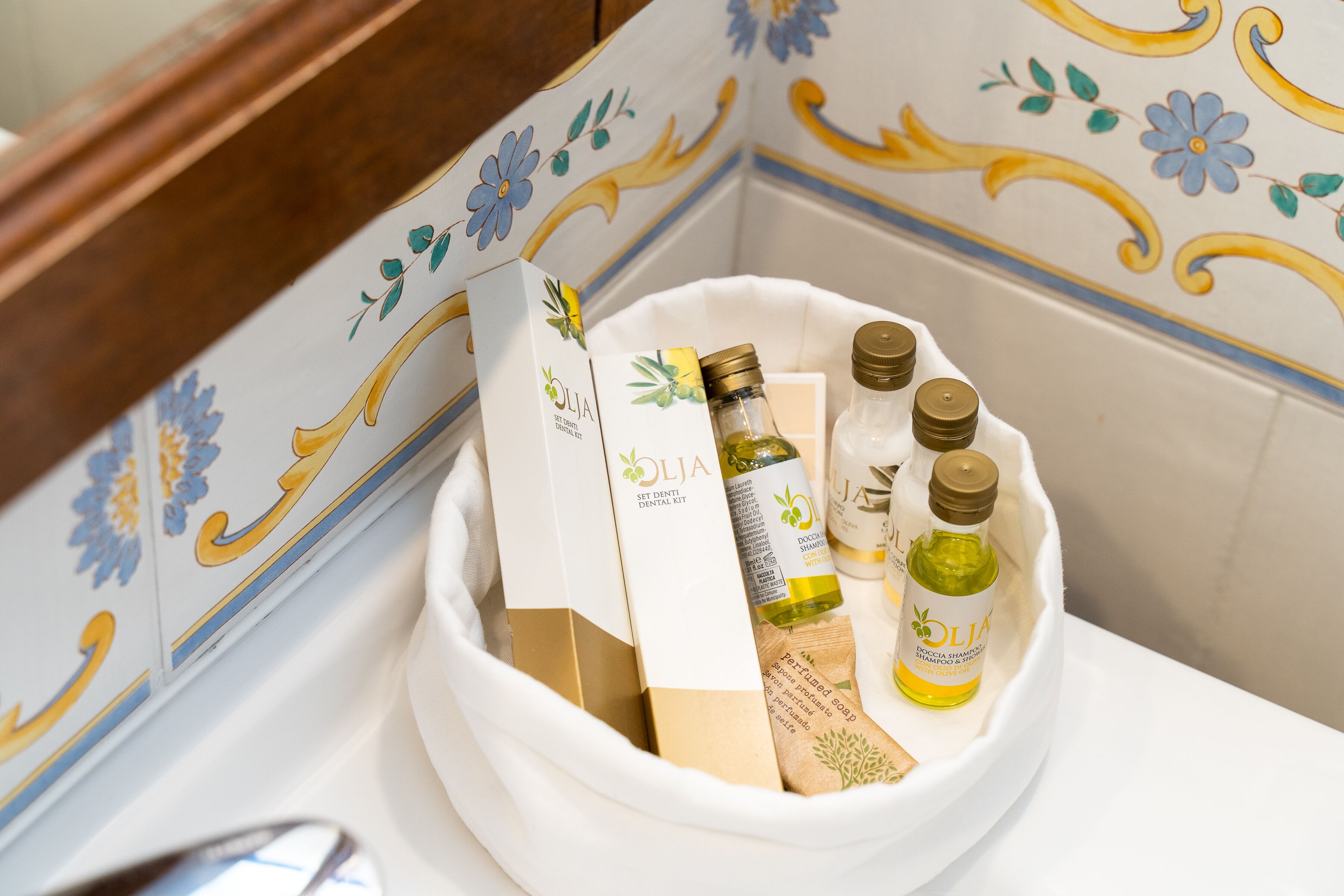 A bathroom with a white sink, a floral tile backsplash, and a white basin containing toiletries including a bottle of olive oil, a bottle of shampoo, and a bar of soap.