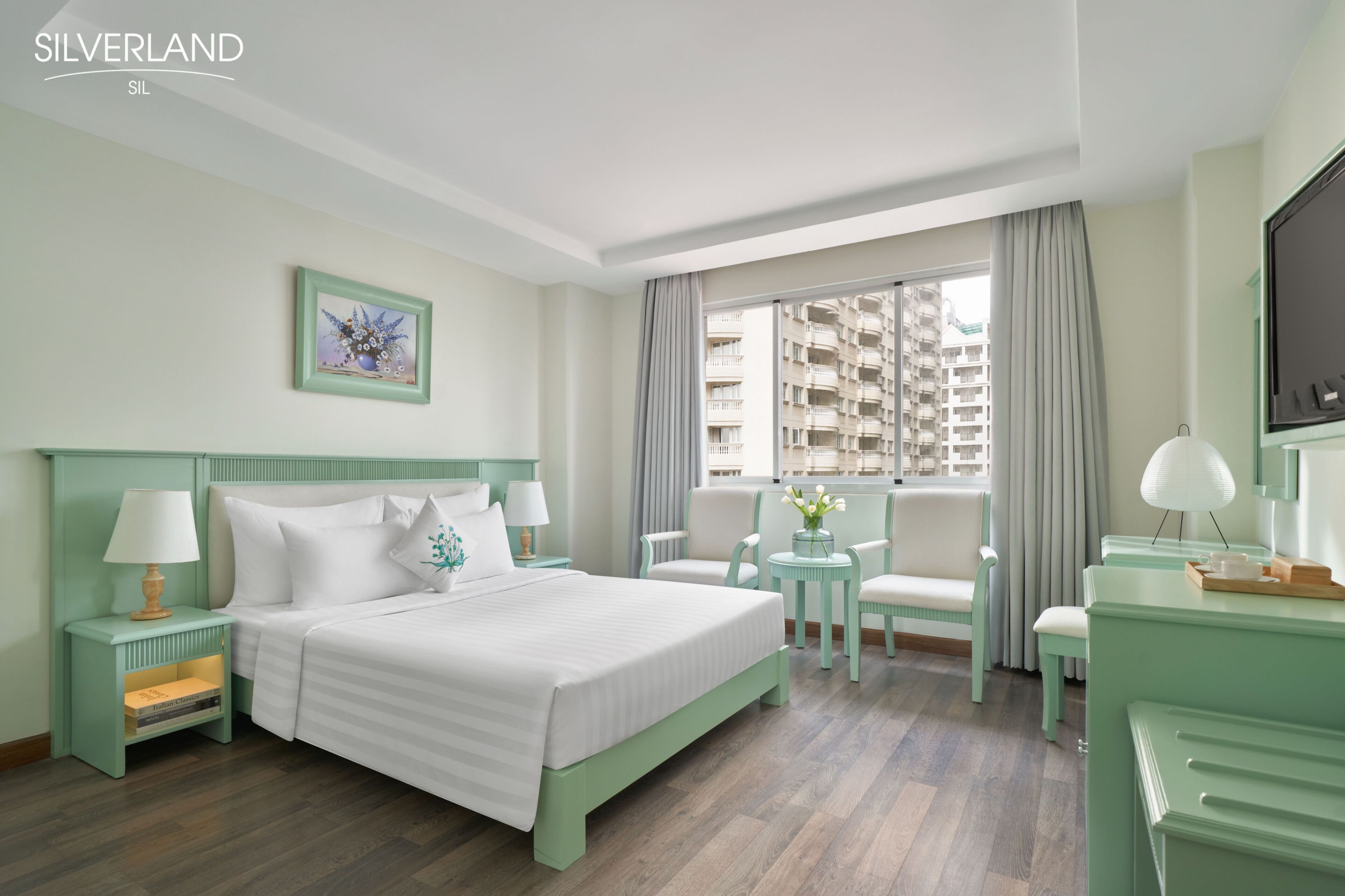 A hotel room with a bed, two nightstands, a desk, a chair, a small table, a TV, and a window with a view of high-rise buildings.