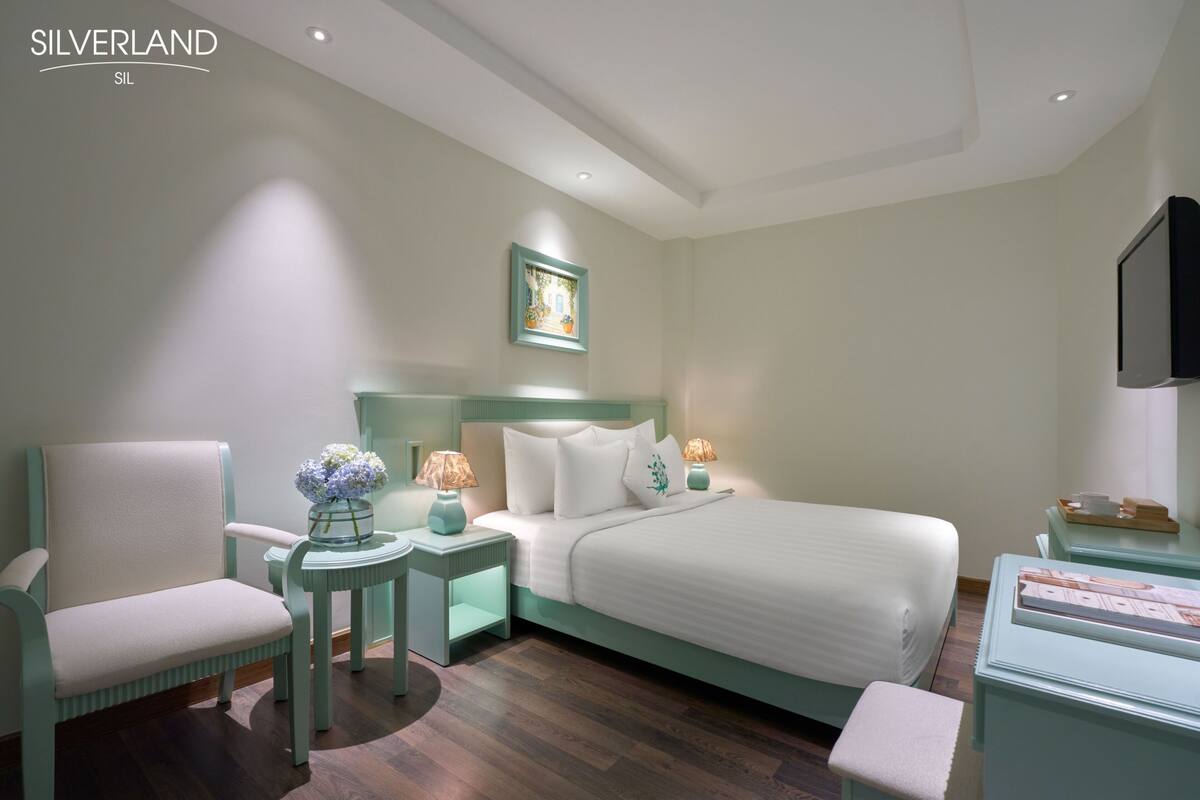 Habitación de hotel con una cama, una silla, una mesita de noche, un televisor y un cuadro en la pared.