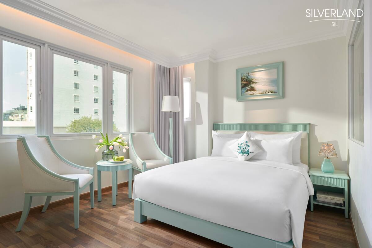 Una habitación de hotel con una cama grande, dos sillas, una mesita con flores, una mesita de noche con lámpara y un cuadro en la pared.