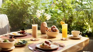 Une table à manger dressée avec différents plats, un grand verre de jus d’orange et un vase rempli de fleurs.