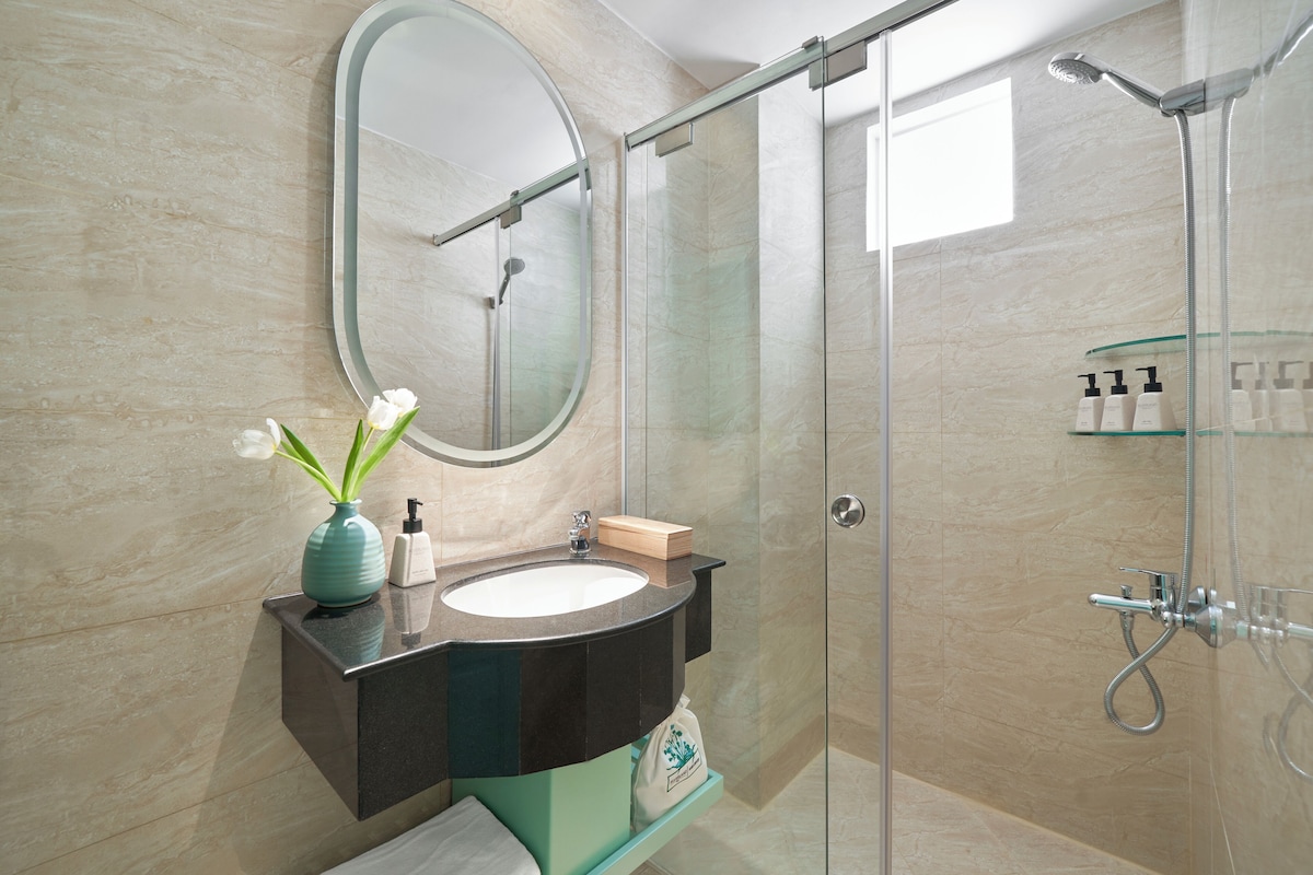 A modern bathroom with a glass-enclosed shower, a round mirror, a sink with a vase of flowers, and a soap dispenser.