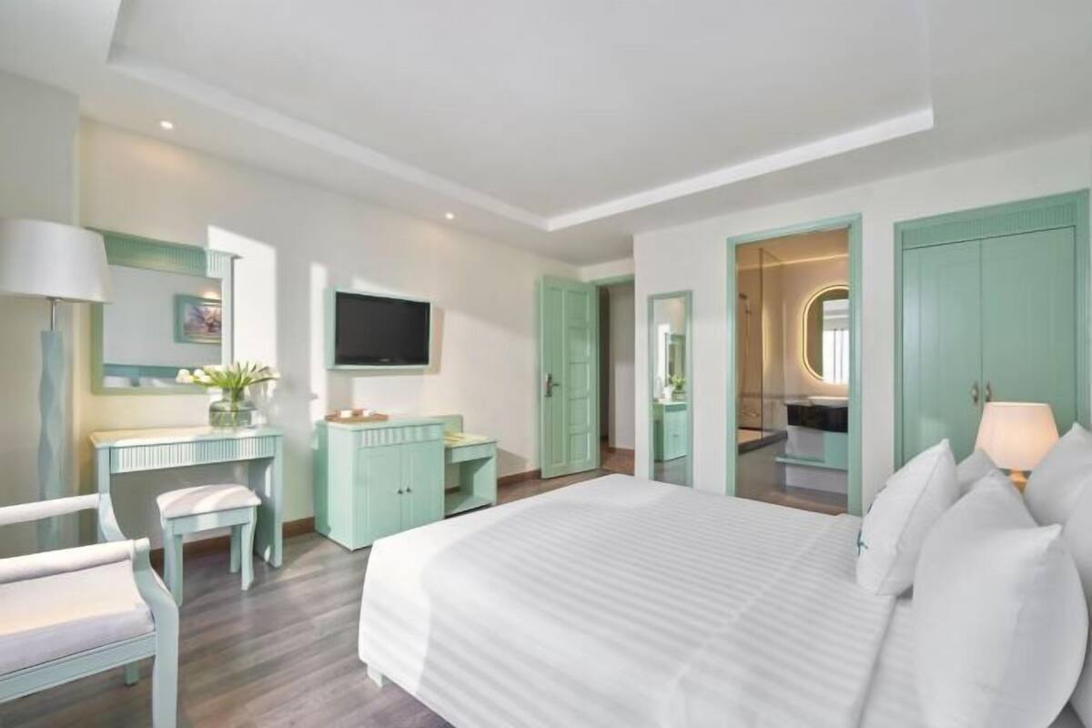 Un dormitorio con una cama grande, un escritorio, un televisor y un baño visible a través de una puerta abierta.