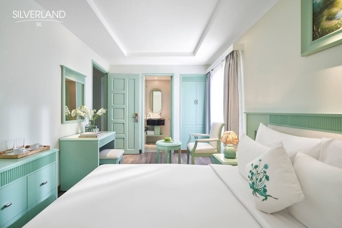 Habitación de hotel con cama, escritorio, silla, mesita pequeña y baño visible a través de una puerta abierta.