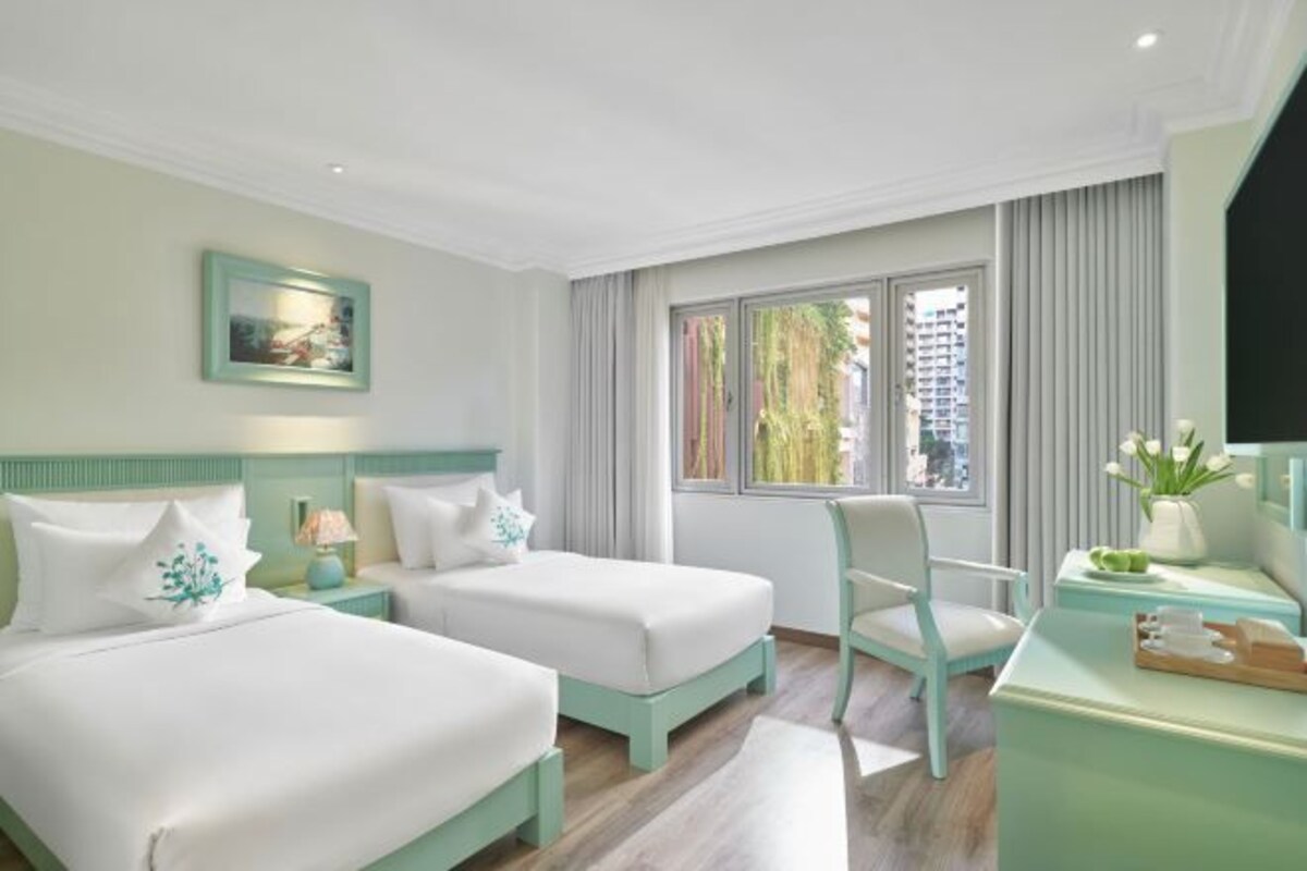 Een hotelkamer met twee bedden, een bureau, een stoel, een raam met gordijnen en een schilderij aan de muur.