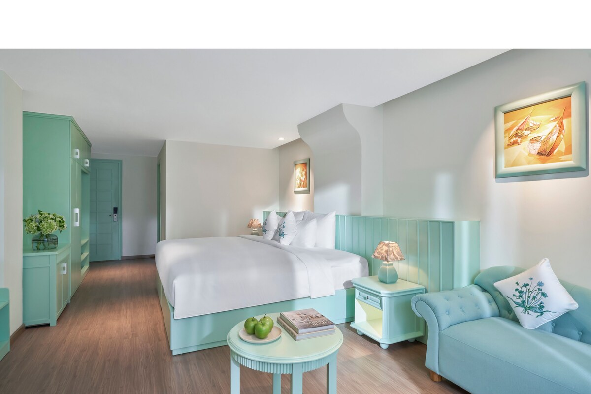 A modern hotel room with a large bed, a small round table with apples and a book, a teal-colored sofa, and a nightstand with a lamp.