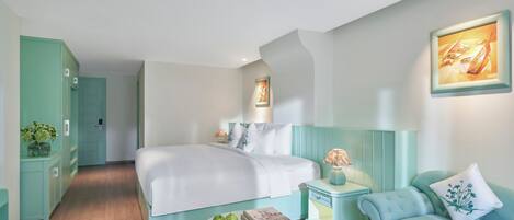 Ein modernes Hotelzimmer mit einem großen Bett, einem kleinen runden Tisch mit Äpfeln und einem Buch, einem türkisfarbenen Sofa und einem Nachttisch mit einer Lampe.